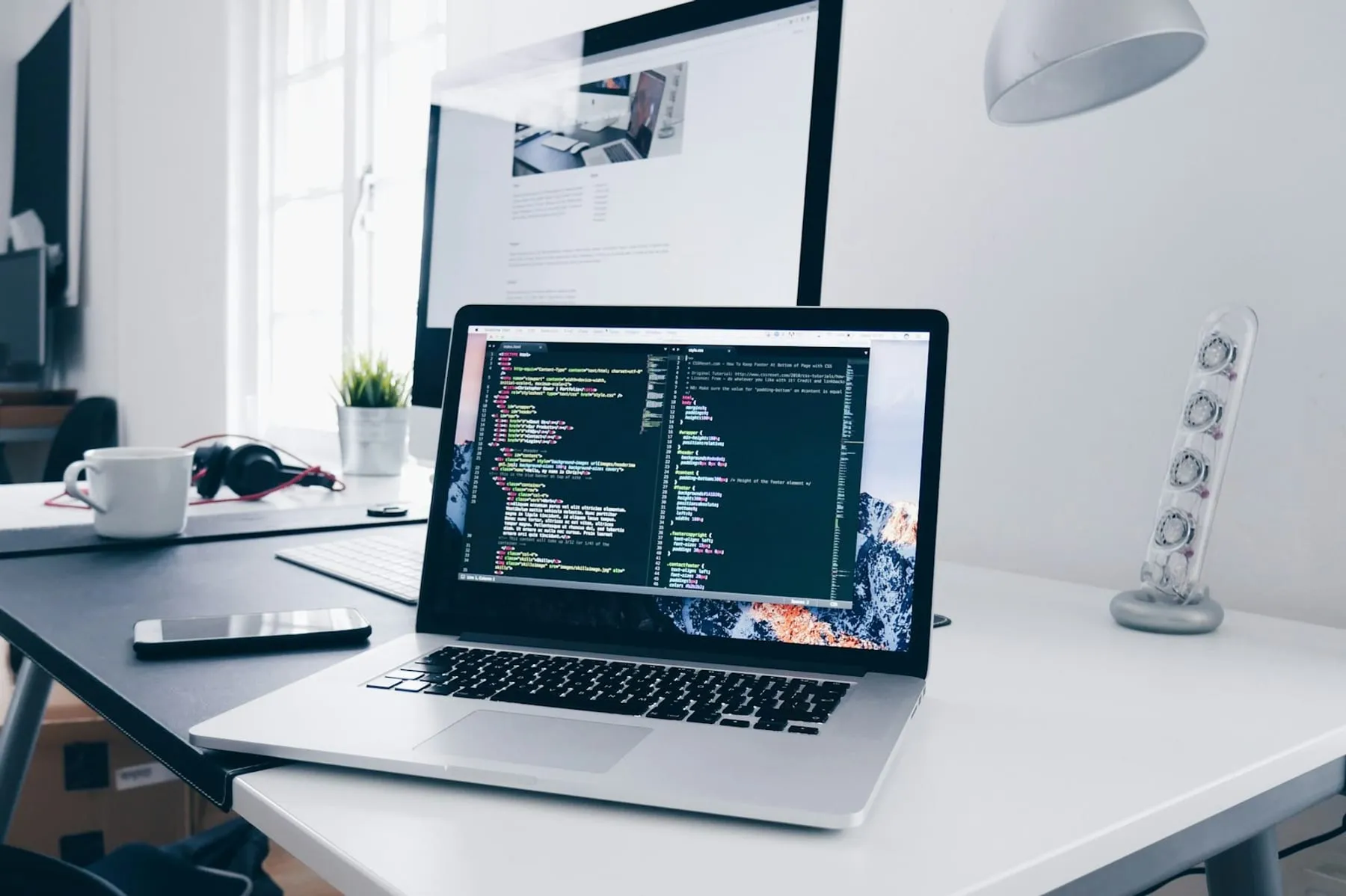 A laptop showing source code on a tidy white desk, beside a coffee cup, a phone and headphones. Nobody is in the picture.